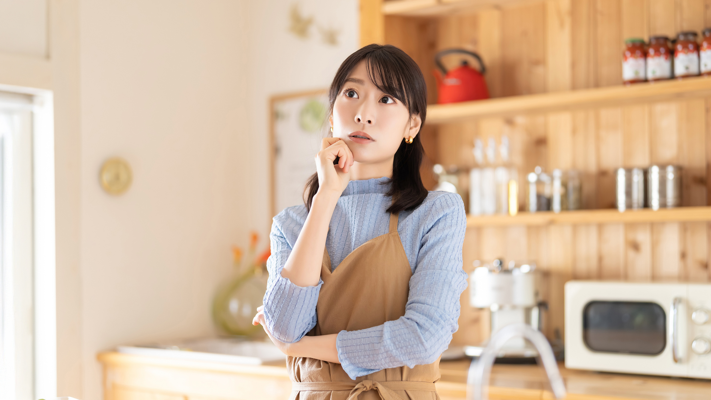 キッチンでエプロン姿の女性が、あごに手を当てて考え込んでいる様子。背後には木製の棚や家電が並び、食事準備について悩んでいる場面。