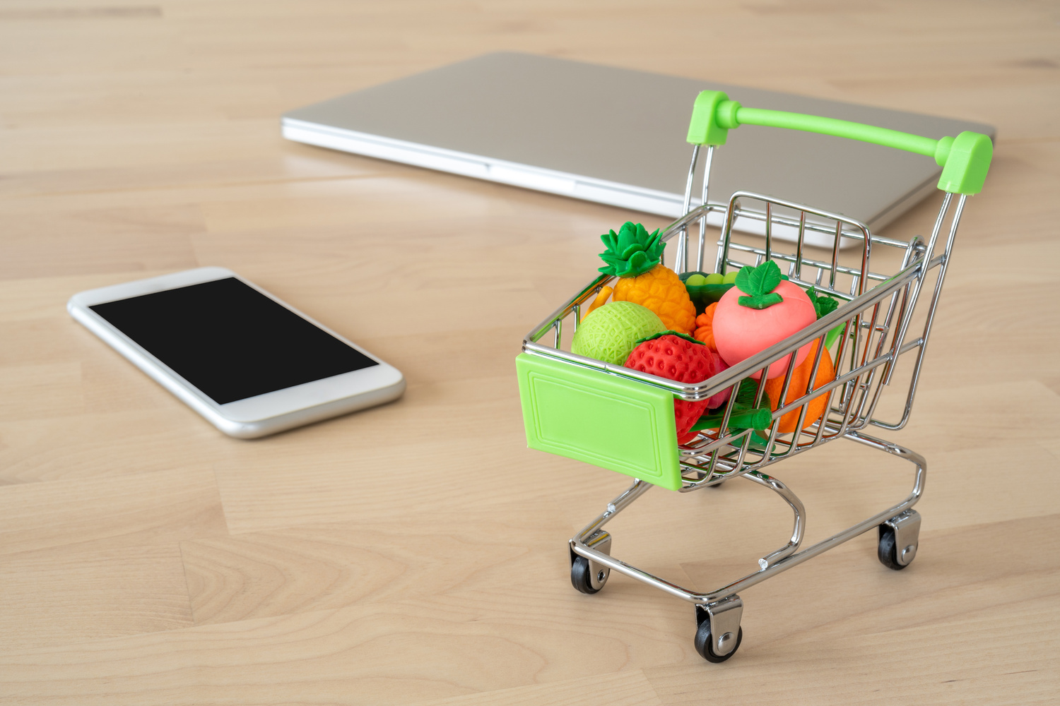 テーブルの上に置かれた小さなショッピングカートとスマートフォン、ノートパソコン。ネットでの買い物やまとめ買い計画をイメージした写真。