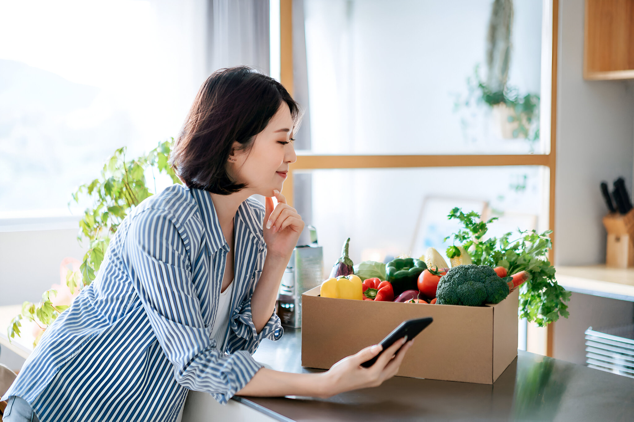 キッチンでスマートフォンを見ながら、野菜の入った宅配ボックスを確認する女性